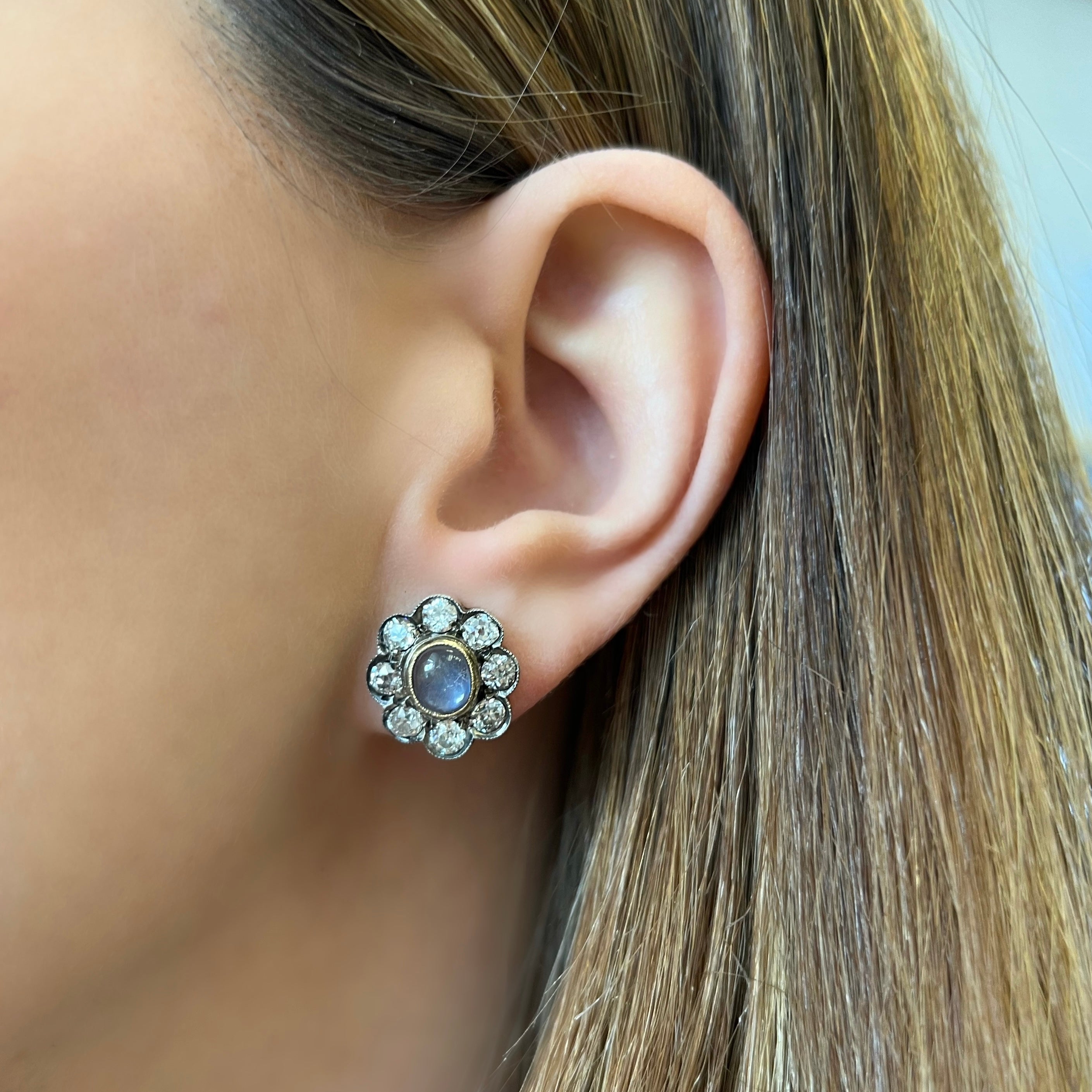 Antique earrings featuring lively bezel-set moonstones, with old mine-cut diamonds in a halo surround, set in silver topped gold. Being worn on the ear.